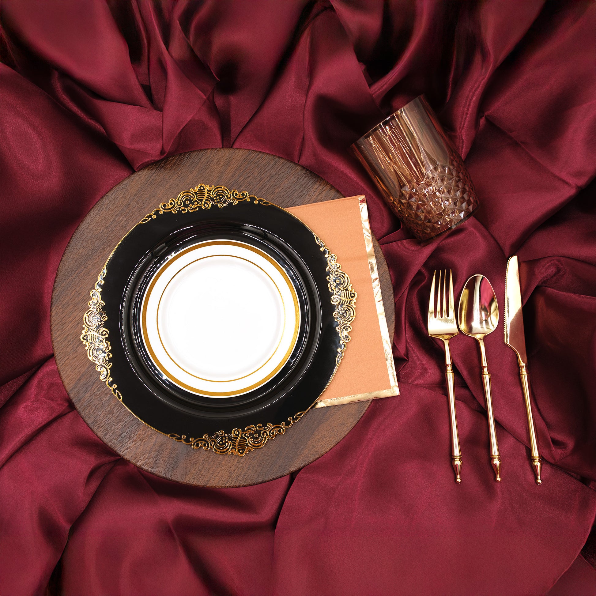 Dinnerware set with a wooden charger plate, black dinner plate, gold cutlery, and amber glass on maroon tablecloth