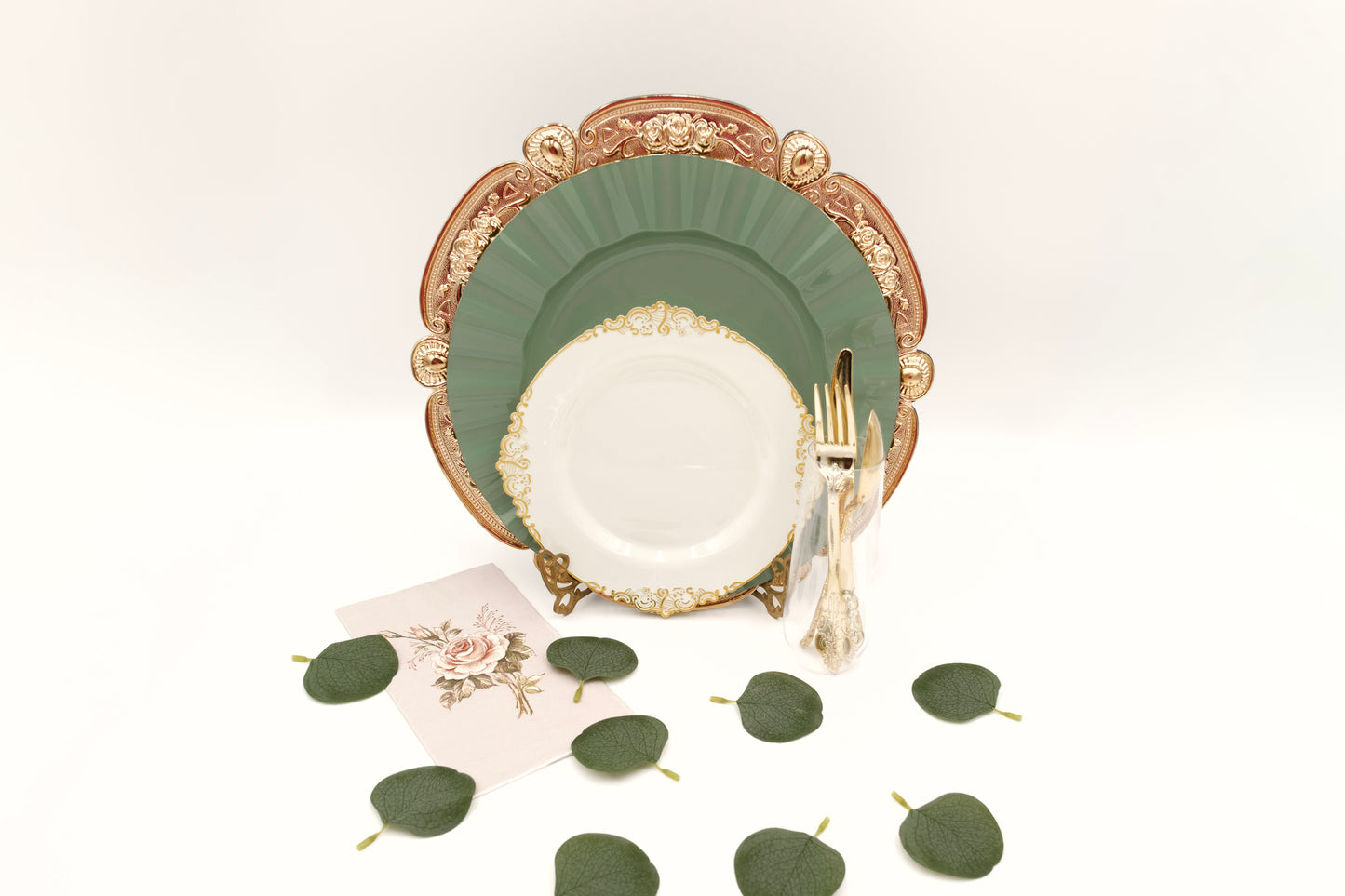 Decorative place setting with gold charger, sage green and ivory plates, gold silverware, and eucalyptus leaves table scatter